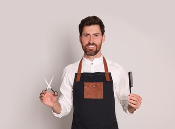 Smiling hairdresser in apron holding scissors and comb on light grey background