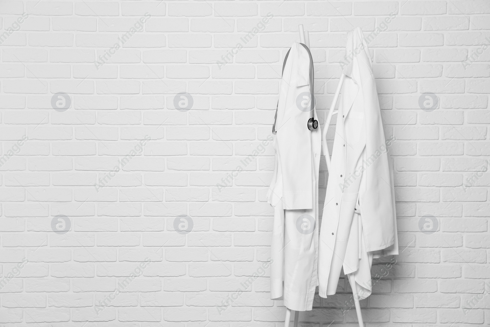 Photo of Medical uniforms and stethoscope hanging on rack near white brick wall. Space for text
