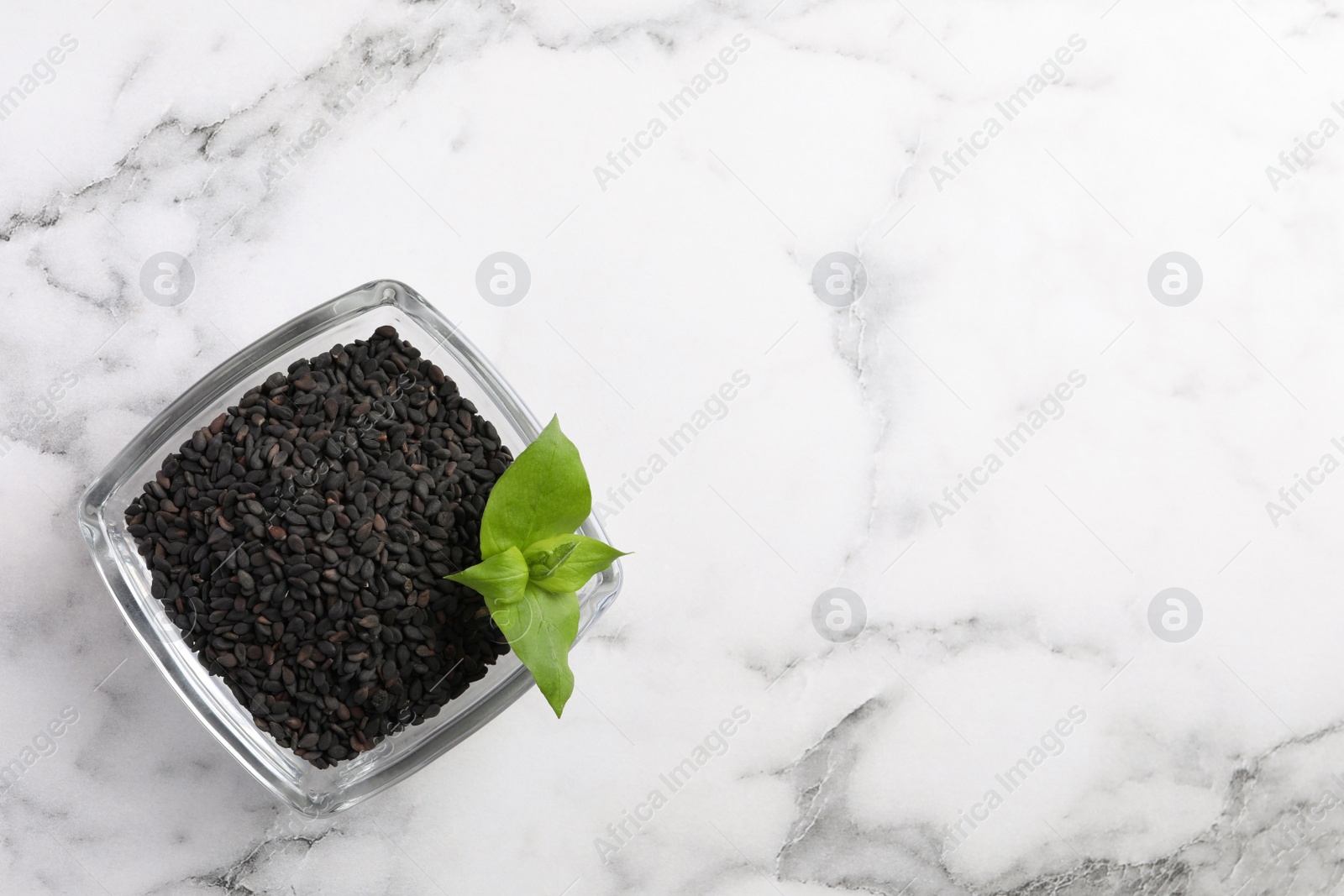 Photo of Black sesame seeds and green leaf in glass bowl on white marble table, top view. Space for text