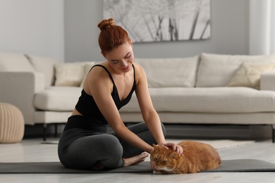 Beautiful woman petting cute red cat on mat at home. Practicing yoga