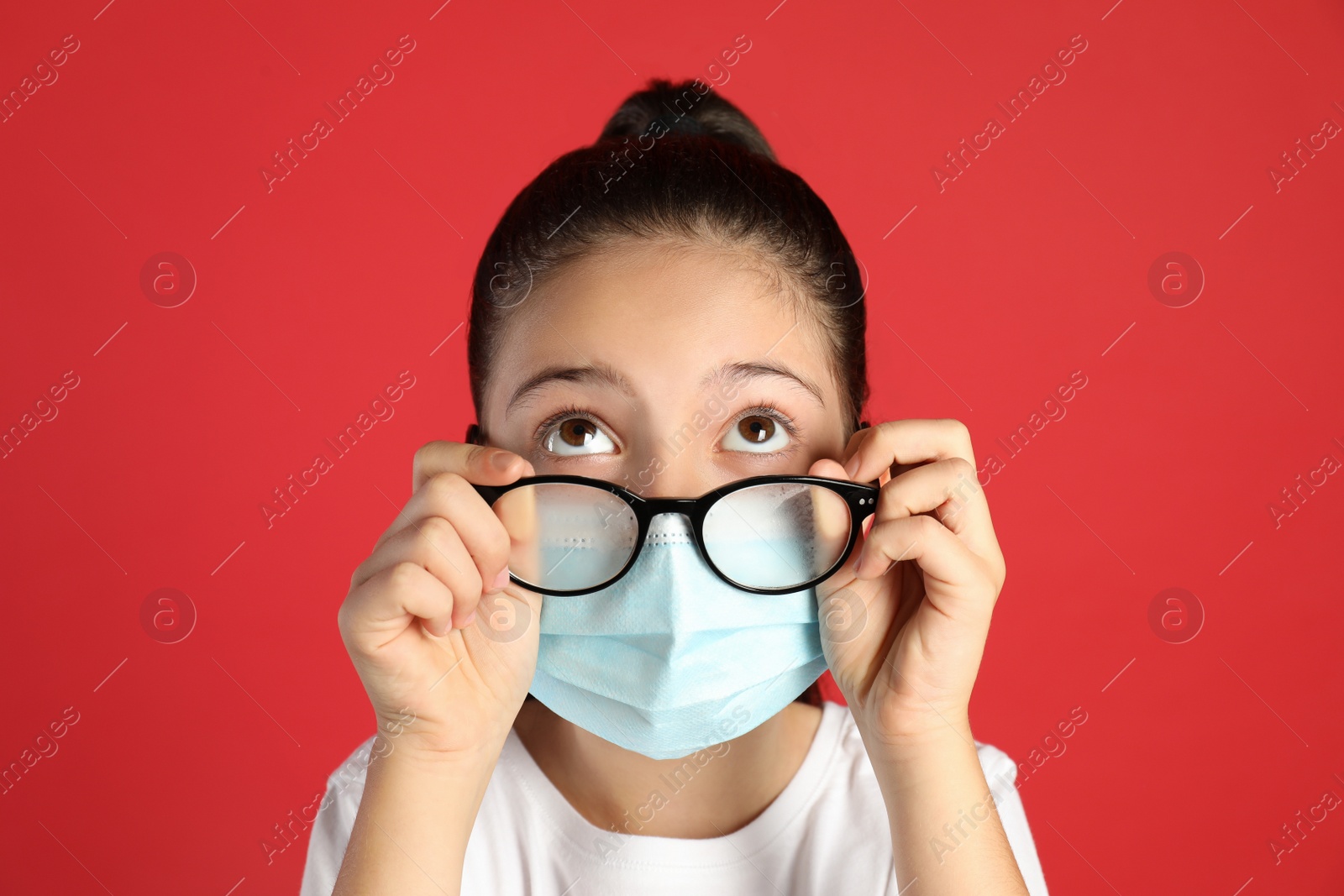 Photo of Little girl wiping foggy glasses caused by wearing medical face mask on red background. Protective measure during coronavirus pandemic