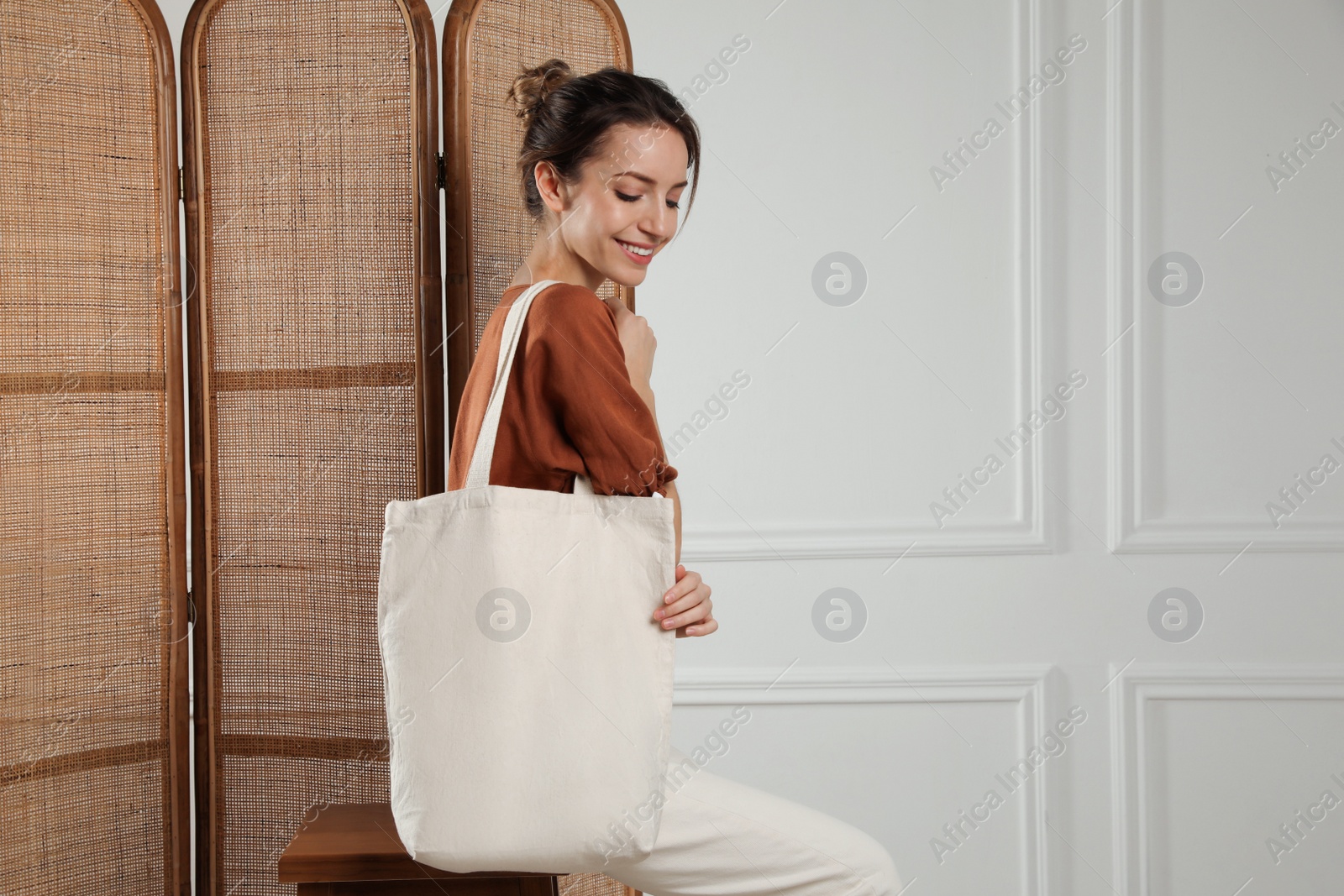 Photo of Happy young woman with blank eco friendly bag sitting on wooden stool indoors. Space for text