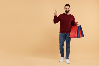 Smiling man with many paper shopping bags showing peace sign on beige background. Space for text