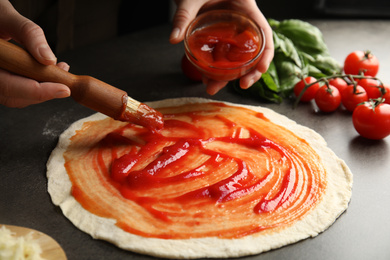 Photo of Woman spreading tomato sauce onto pizza crust at grey table, closeup