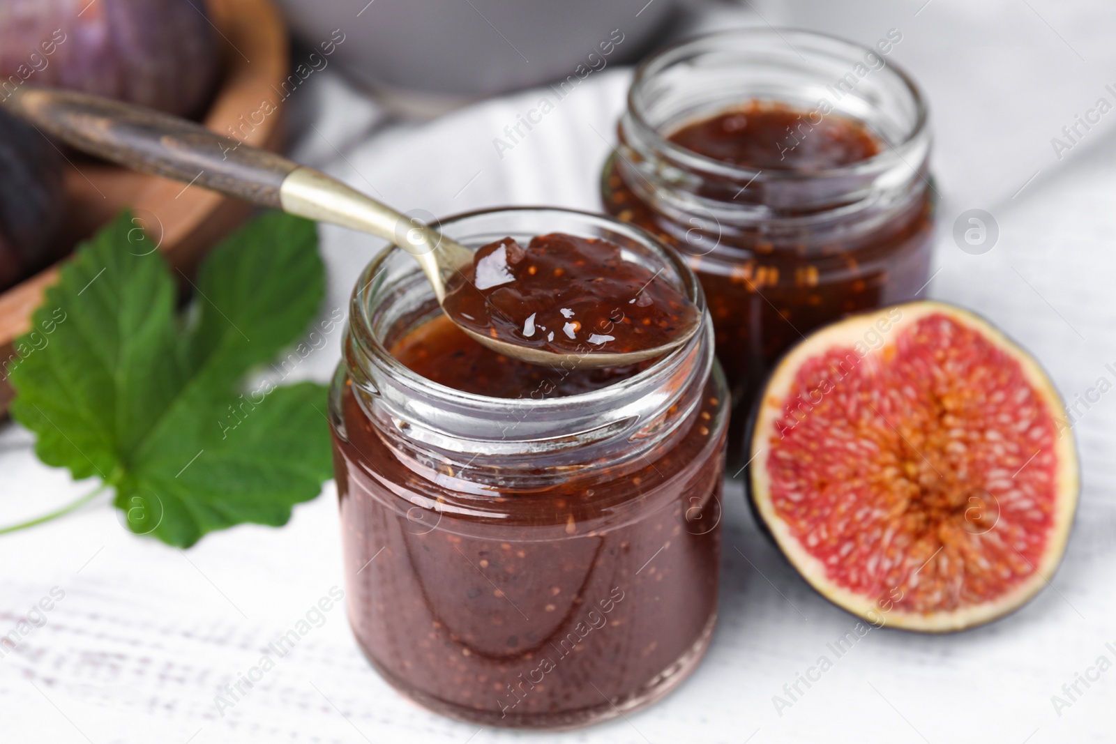 Photo of Glass jars of tasty sweet fig jam with spoon and fruits on white wooden table, closeup