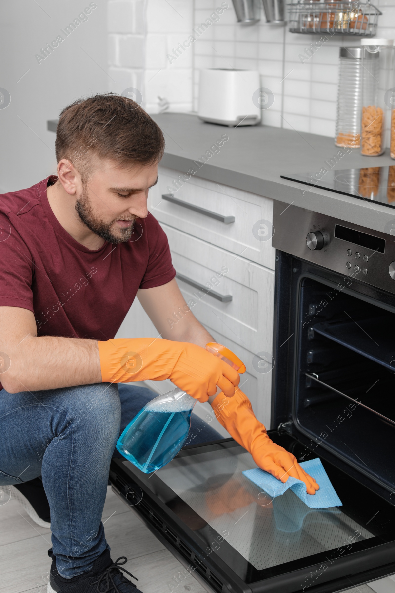 Photo of Young man cleaning oven with rag and detergent in kitchen