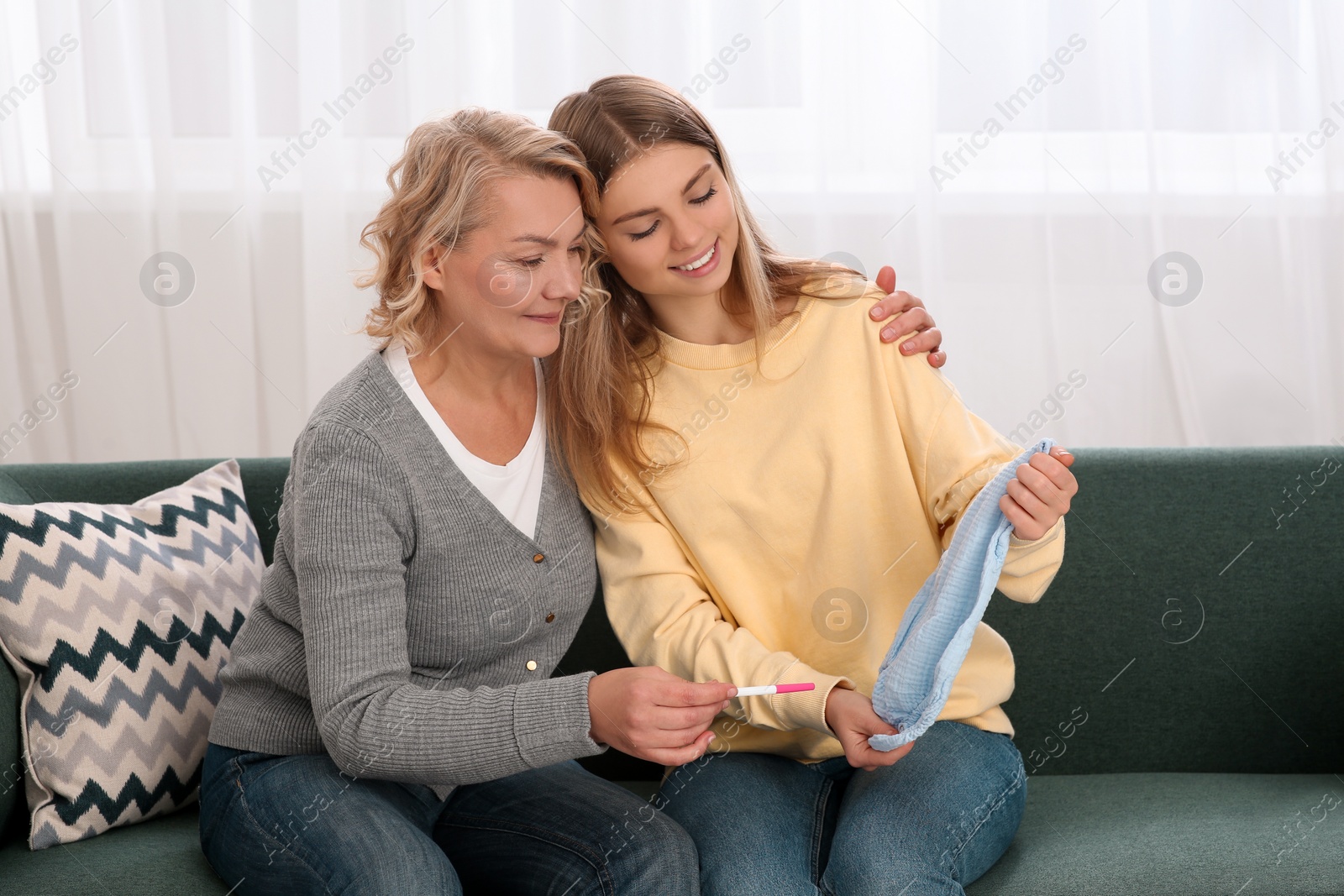 Photo of Young woman showing baby's bodysuit to her mother with pregnancy test at home. Grandparent reaction to future grandson