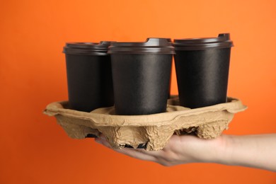 Woman holding cardboard holder with takeaway paper coffee cups against orange background, closeup