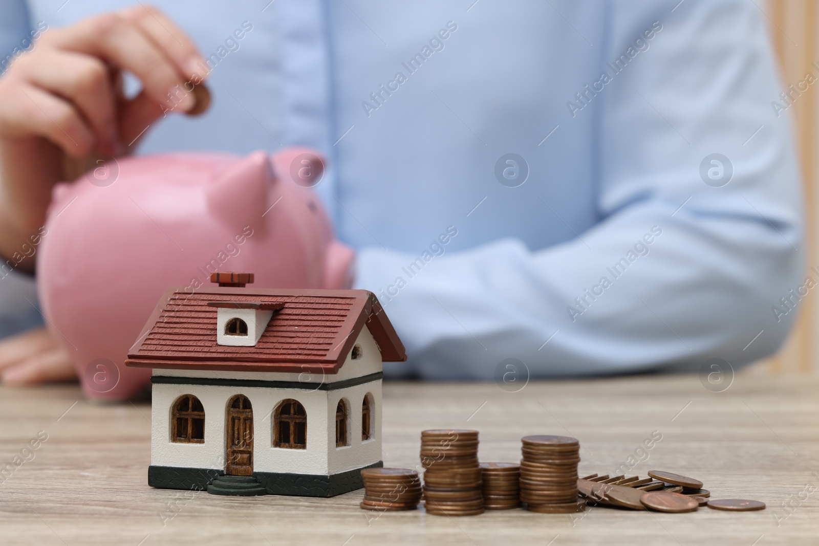 Photo of Woman with piggy bank at wooden table, focus on house model and stacked coins