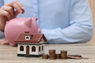 Photo of Woman with piggy bank at wooden table, focus on house model and stacked coins