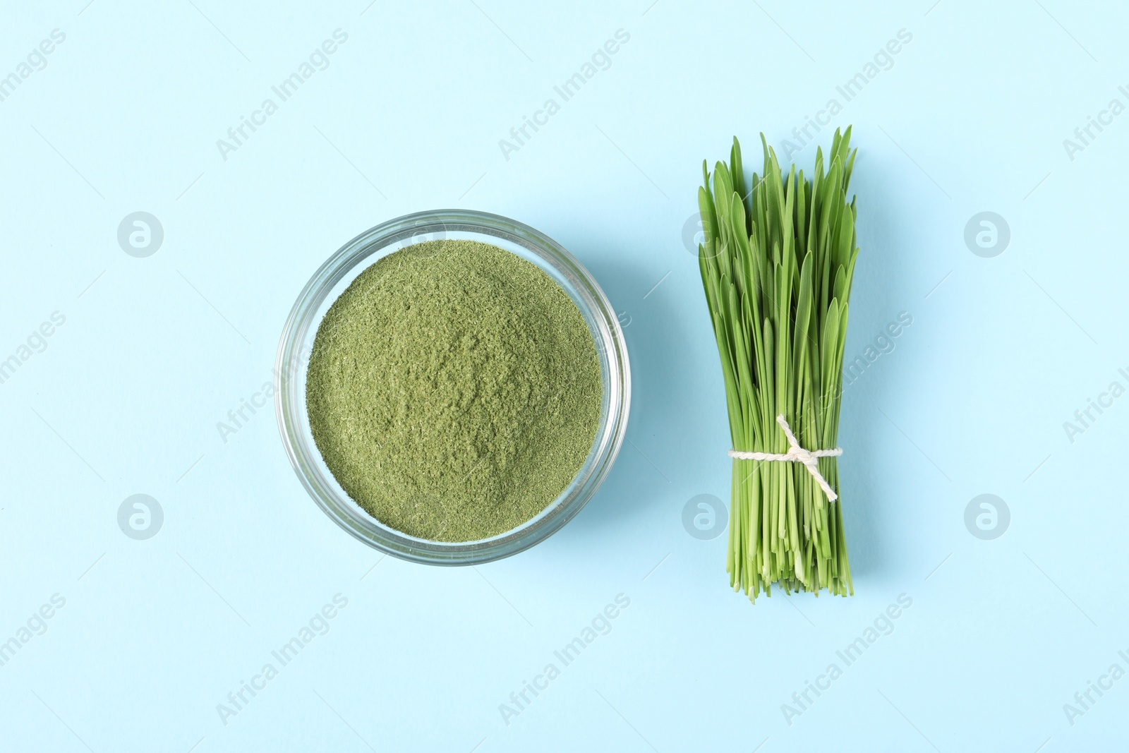 Photo of Wheat grass powder in glass bowl and fresh sprouts on light blue table, flat lay