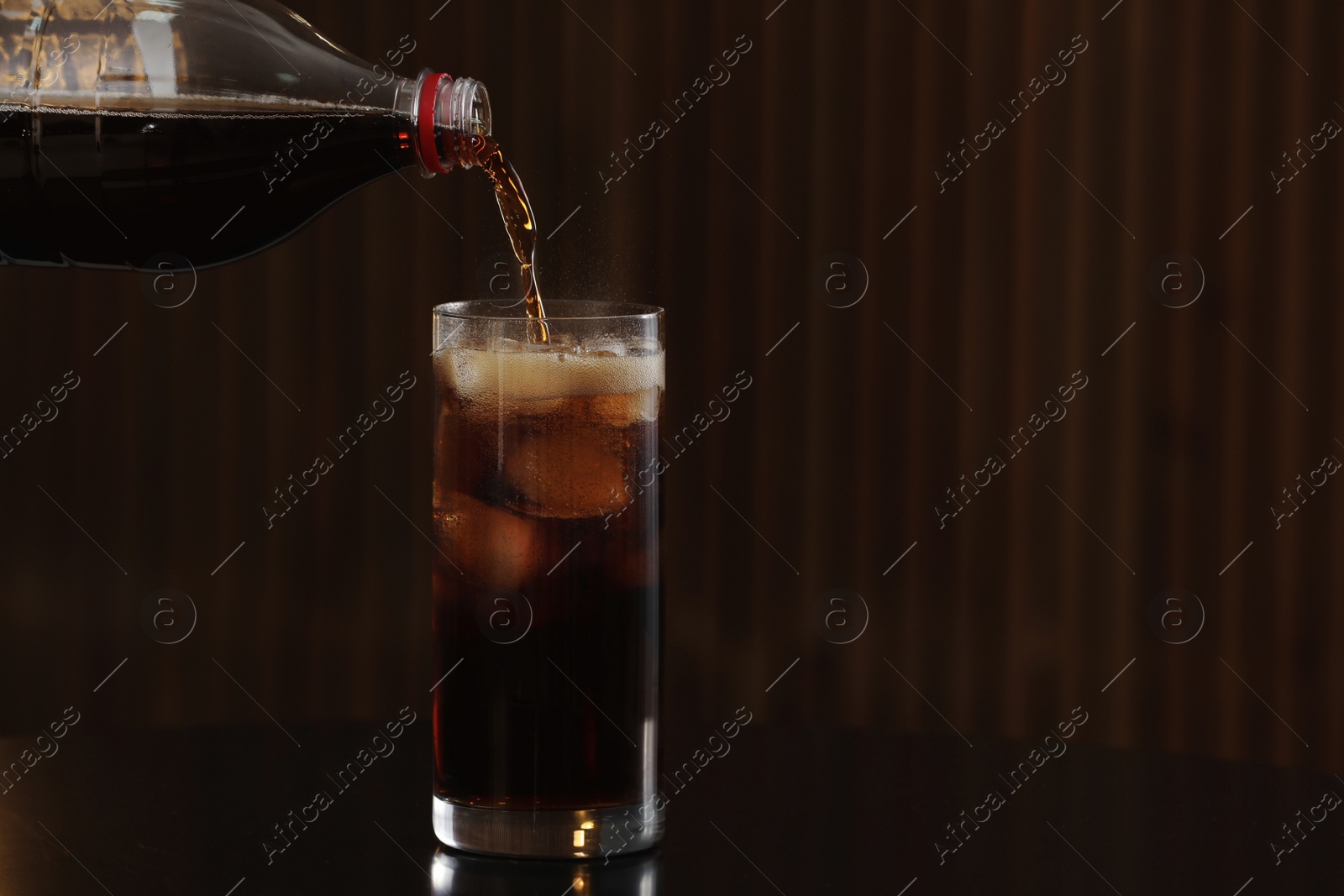 Photo of Pouring cola from bottle into glass with ice cubes on table against blurred background. Space for text