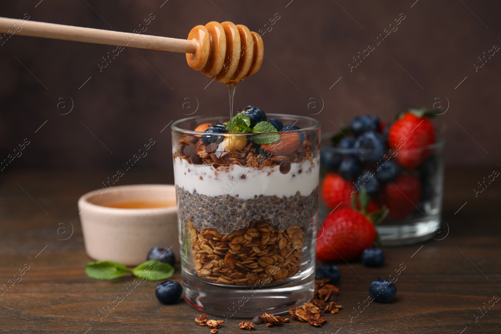 Photo of Pouring honey onto tasty granola with berries, yogurt and chia seeds at wooden table