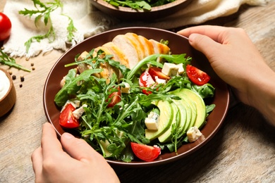 Woman holding bowl of fresh salad with arugula and chicken at wooden table, closeup