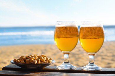 Glasses of cold beer and pistachios on wooden table near sea, space for text