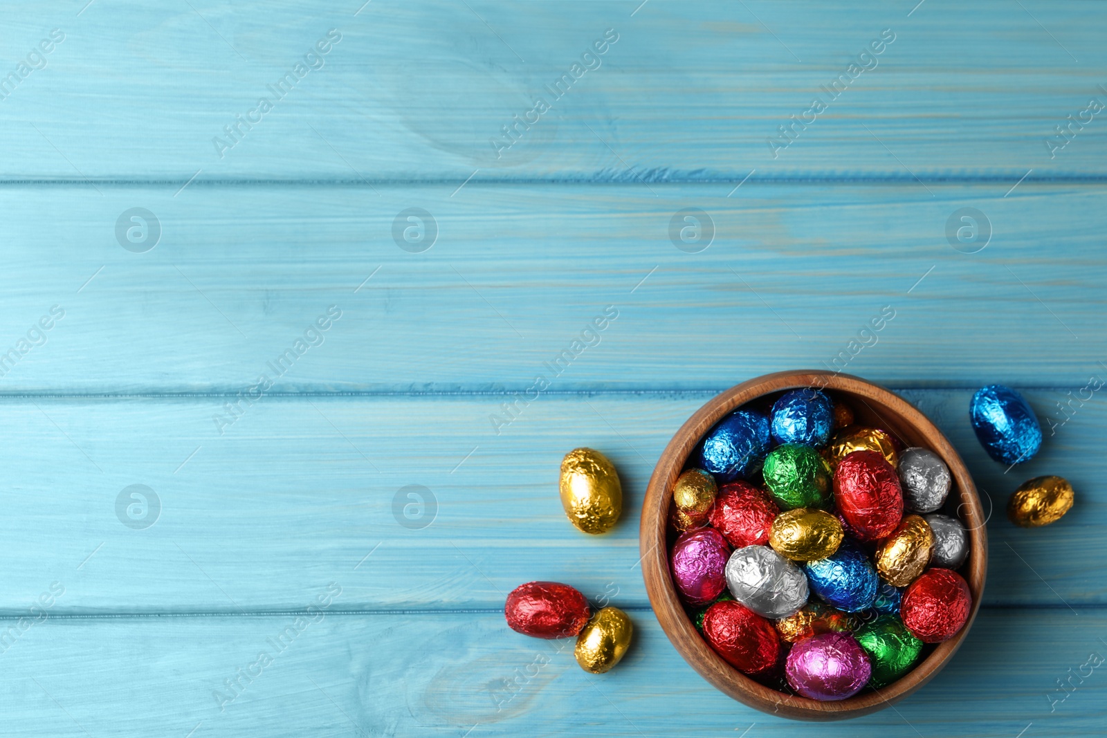 Photo of Bowl with chocolate eggs wrapped in colorful foil on light blue wooden table, flat lay. Space for text