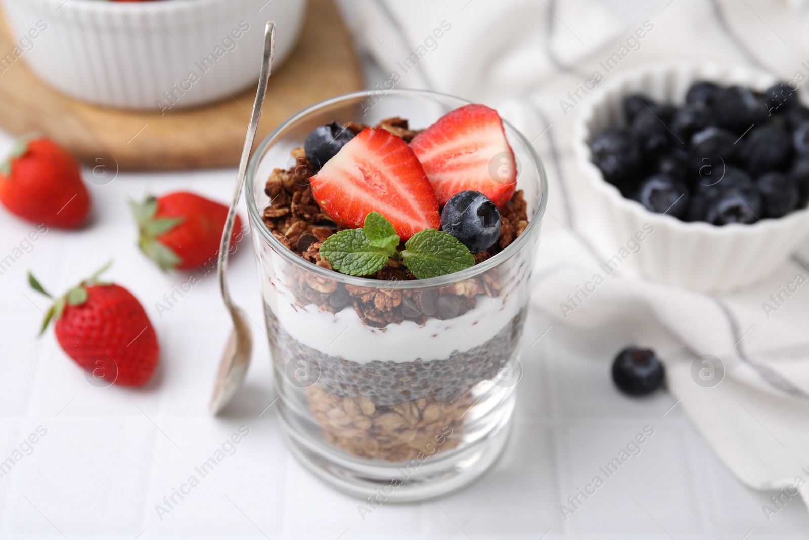 Photo of Tasty granola with berries, yogurt and chia seeds in glass on white tiled table, closeup