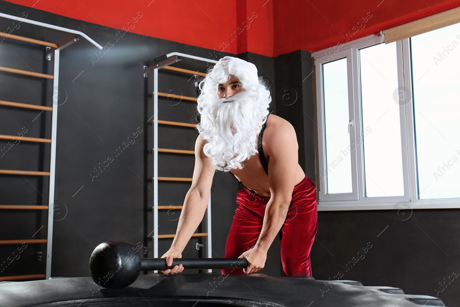 Photo of Young Santa Claus hitting heavy tire with hammer in modern gym