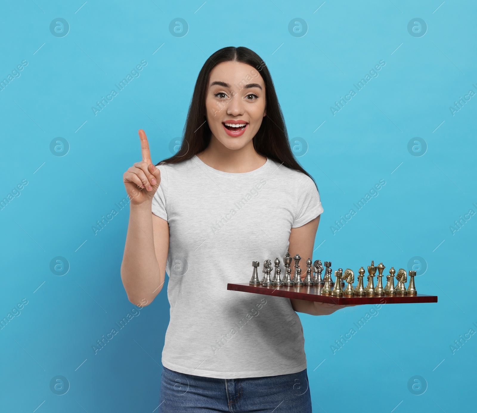 Photo of Happy woman with chessboard pointing upwards on light blue background