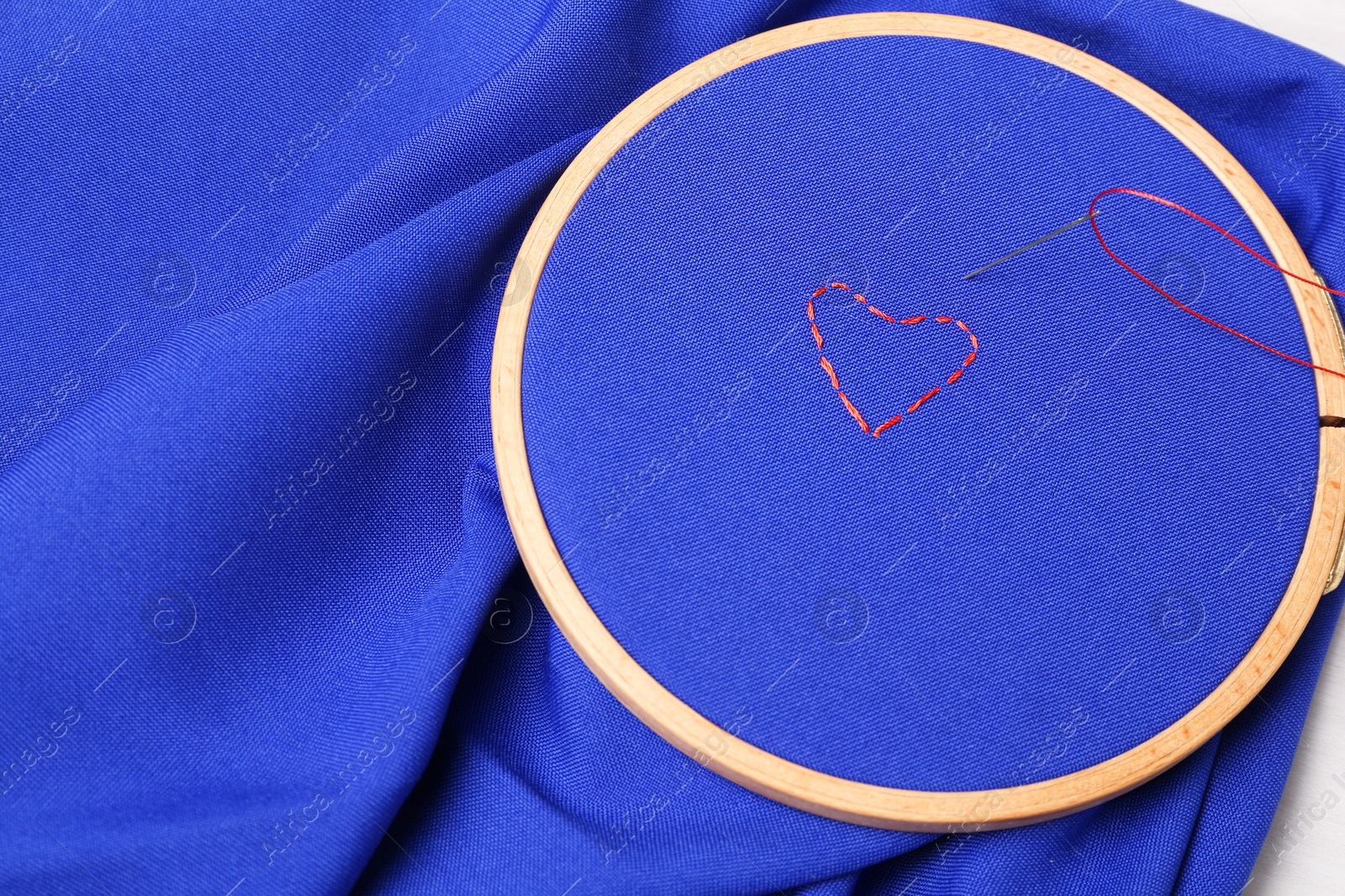 Photo of Red embroidered heart, thread and sewing needle on blue cloth with hoop, above view