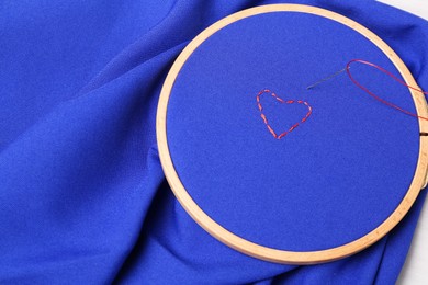Red embroidered heart, thread and sewing needle on blue cloth with hoop, above view