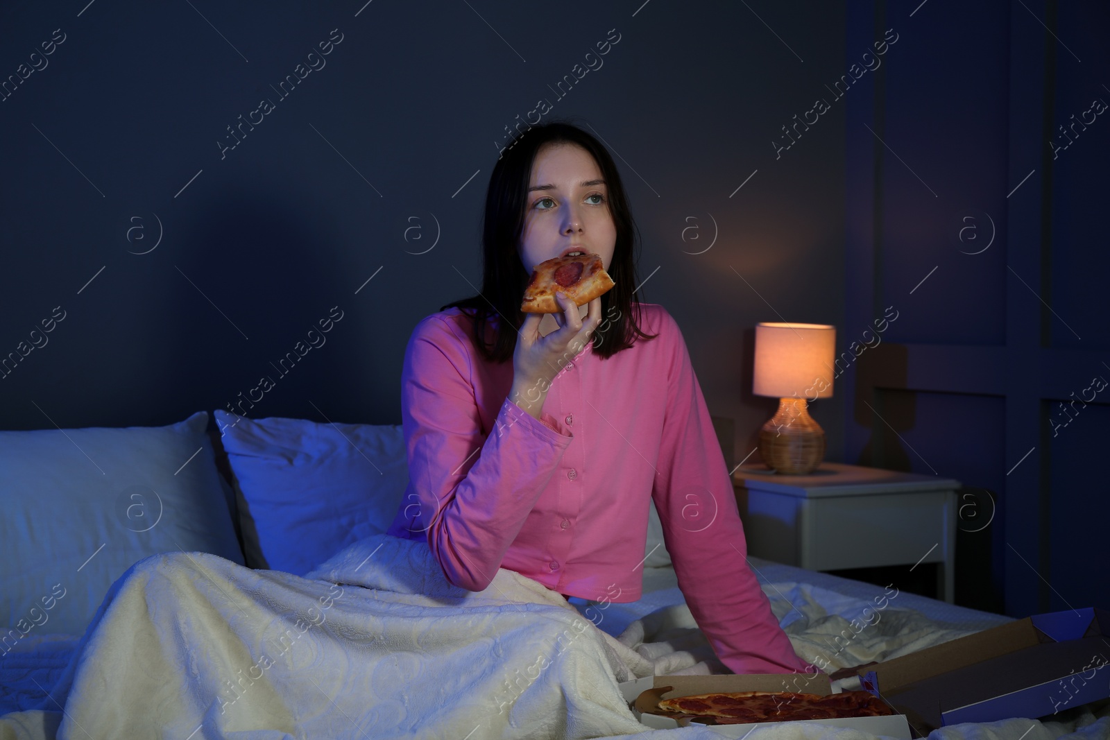 Photo of Young woman eating pizza while watching TV on bed at night