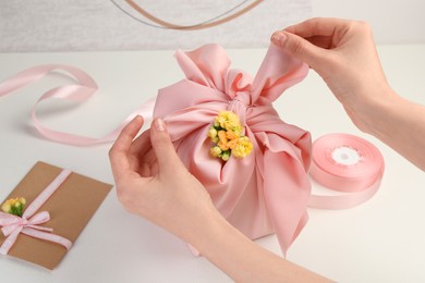 Photo of Furoshiki technique. Woman wrapping gift in pink fabric with flowers at white table, closeup