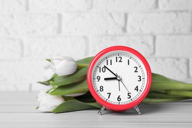 Photo of Red alarm clock and beautiful tulips on white wooden table against brick wall. Spring time