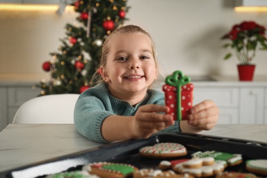 Photo of Cute little girl taking fresh Christmas gingerbread cookie from baking sheet at table indoors