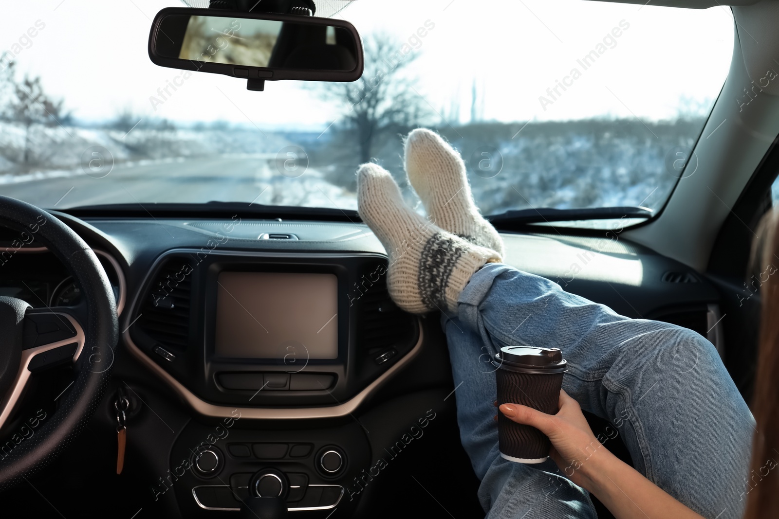 Photo of Young woman in warm socks holding her legs on car dashboard and drinking coffee. Cozy atmosphere