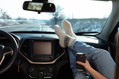 Photo of Young woman in warm socks holding her legs on car dashboard and drinking coffee. Cozy atmosphere