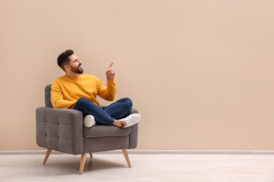 Photo of Handsome man pointing at something while sitting in armchair near beige wall indoors, space for text