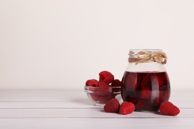 Photo of Jar of tasty canned raspberry jam and fresh berries in glass bowl on white wooden table, space for text