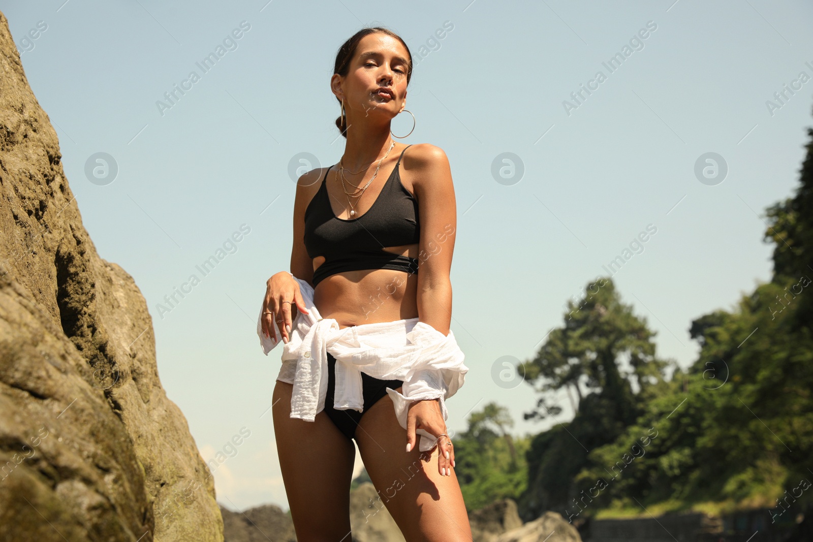 Photo of Beautiful young woman in stylish bikini near big stone outdoors on sunny day, low angle view