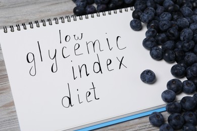Photo of Notebook with words Low Glycemic Index Diet and blueberries on light wooden table, closeup