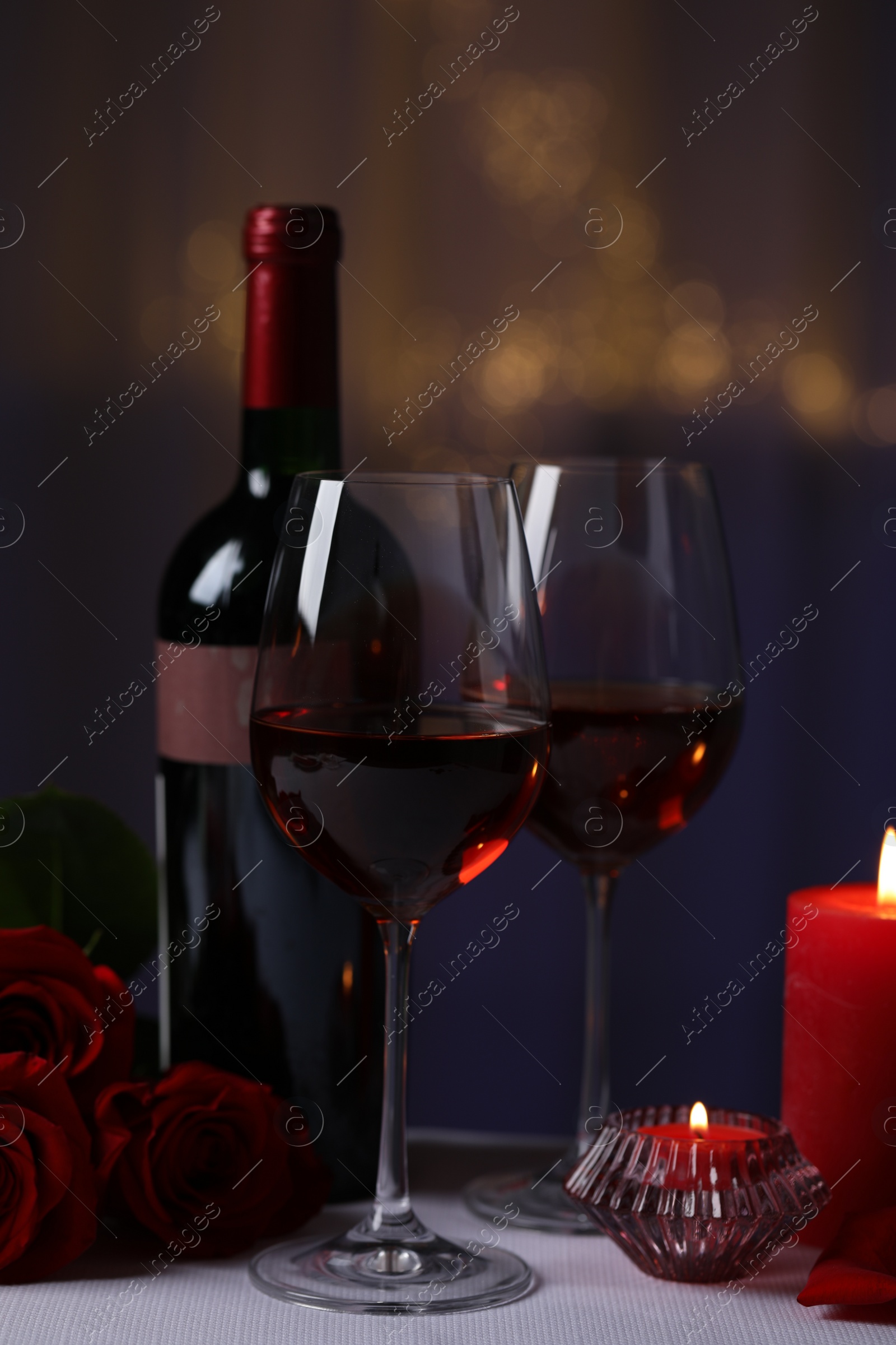 Photo of Glasses of red wine, rose flowers and burning candles on grey table against blurred lights. Romantic atmosphere