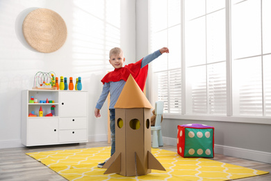 Little child in red cape playing with rocket made of cardboard box at home