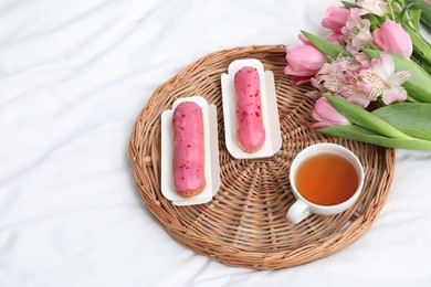 Photo of Tasty breakfast served in bed. Delicious eclairs, tea and flowers on wicker tray, above view with space for text