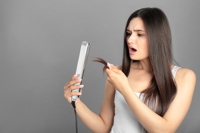 Upset woman with hair iron on grey background, space for text