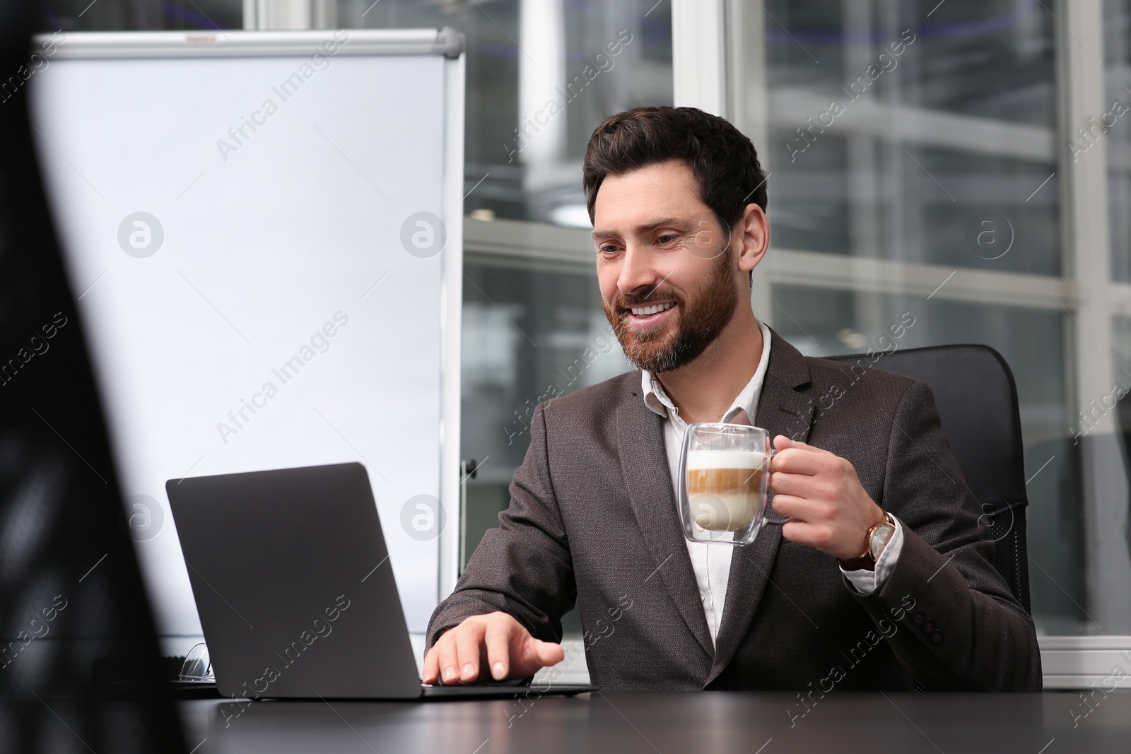 Photo of Man with cup of coffee working on laptop at black desk in office