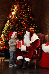 Santa Claus and little boy with gift near Christmas tree indoors
