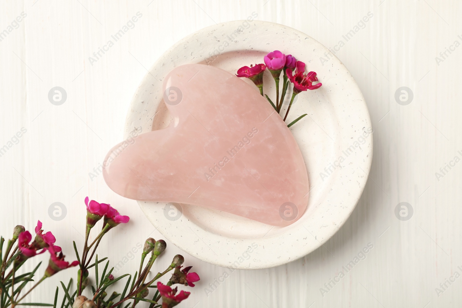 Photo of Rose quartz gua sha tool and flowers on white wooden table, flat lay