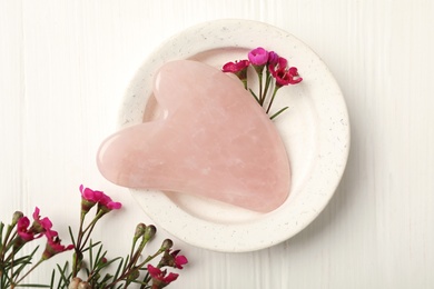 Photo of Rose quartz gua sha tool and flowers on white wooden table, flat lay
