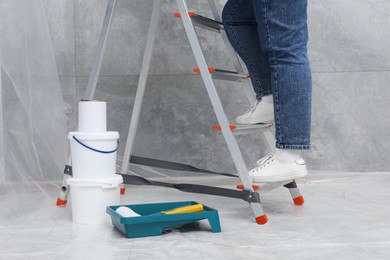 Photo of Woman climbing metallic folding ladder, tray with roller and buckets of paint indoors, closeup
