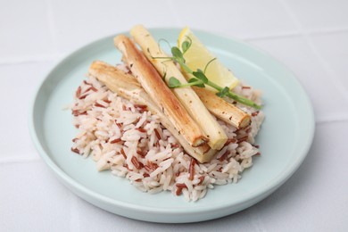 Photo of Plate with baked salsify roots, lemon and rice on white tiled table