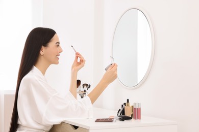 Photo of Beautiful young woman applying eyeshadow with brush at dressing table indoors