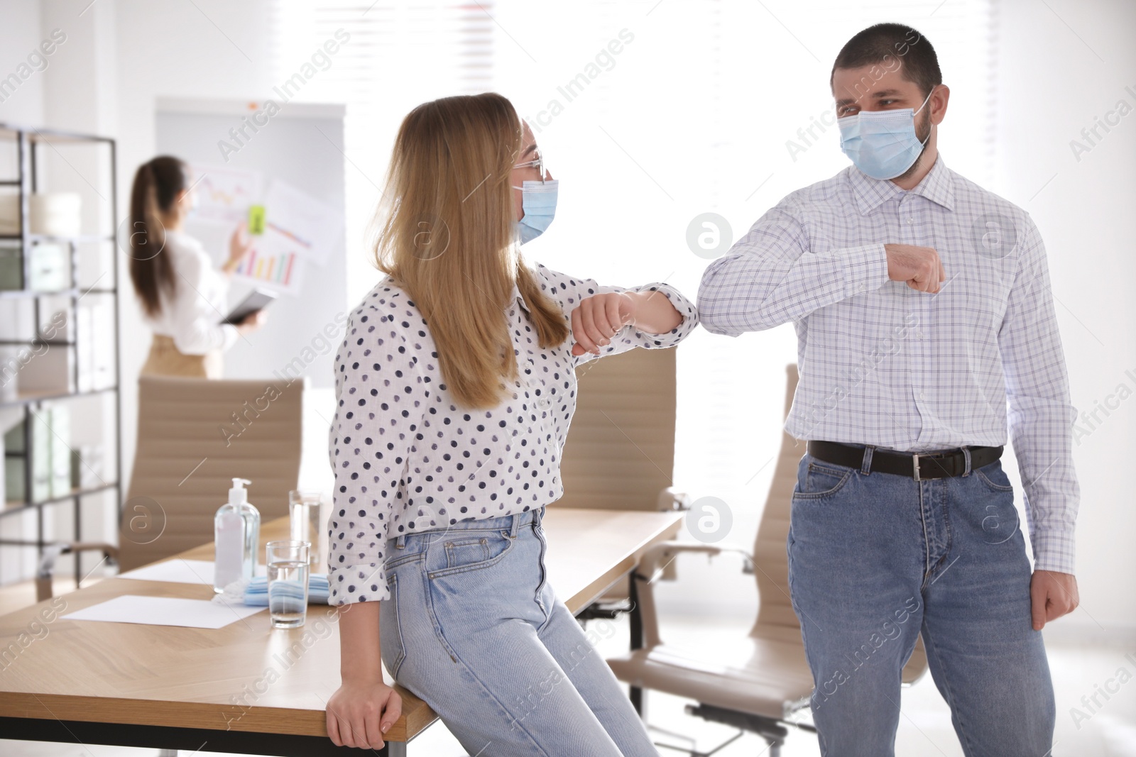 Photo of Coworkers with protective masks making elbow bump in office. Informal greeting during COVID-19 pandemic