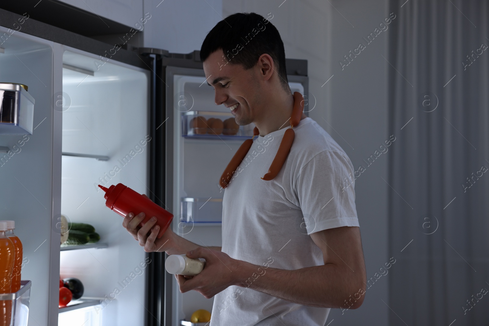 Photo of Happy man with sausages and sauces near refrigerator in kitchen at night