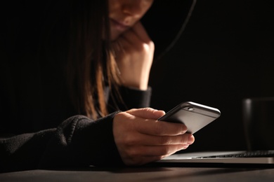 Woman using smartphone at table with laptop in darkness, closeup. Loneliness concept