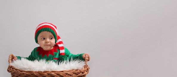Cute baby wearing elf costume near wicker basket on light grey background, banner design with space for text. Christmas celebration